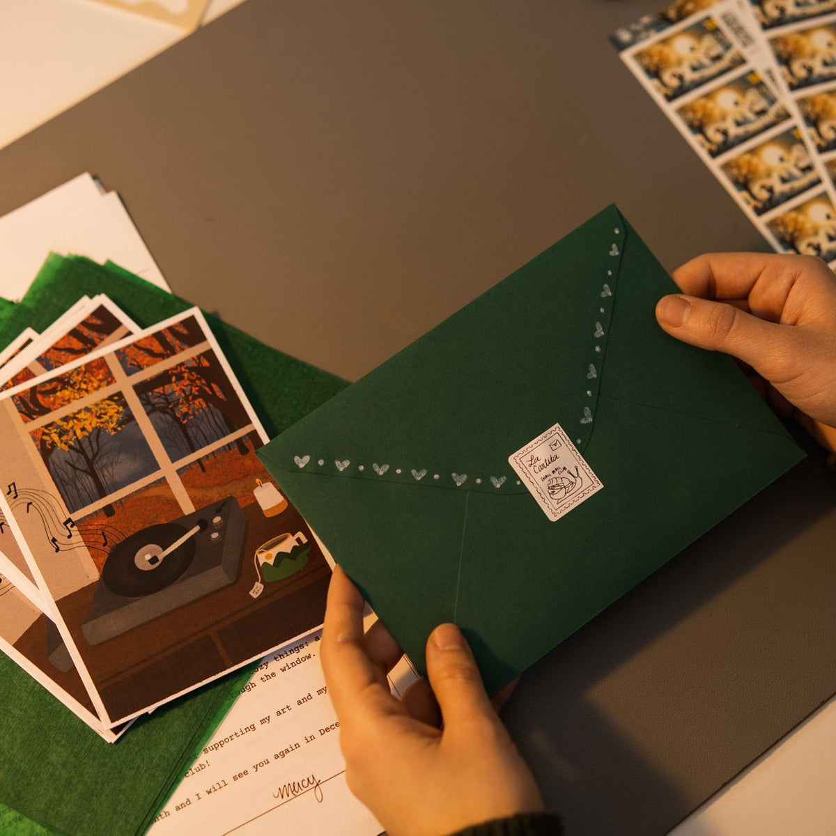 Person holding a green envelope with a stamp on a table with postcards and stamps.