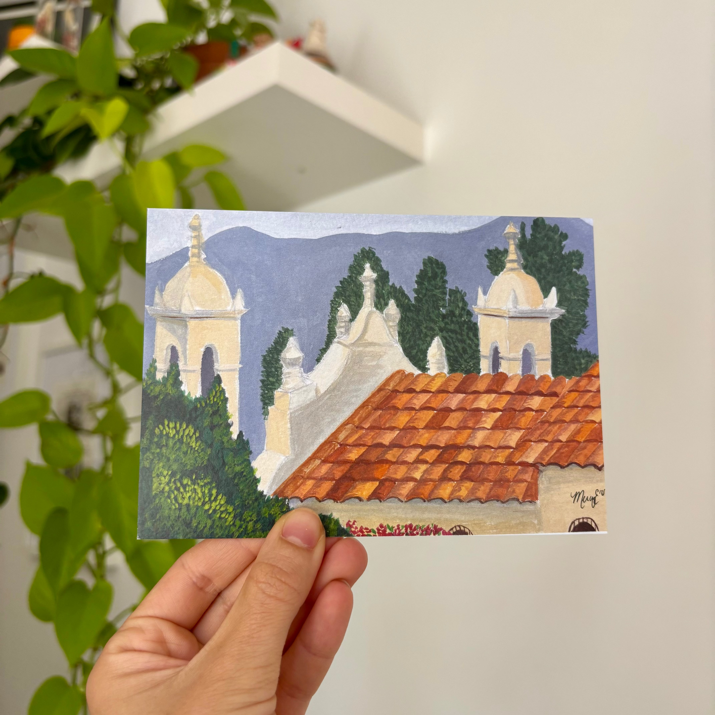 Hand holding a small painting of a scenic landscape with buildings and trees.