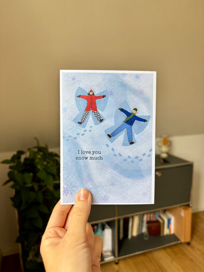 Hand holding a card with two people snow angels and 'I love you snow much' text.